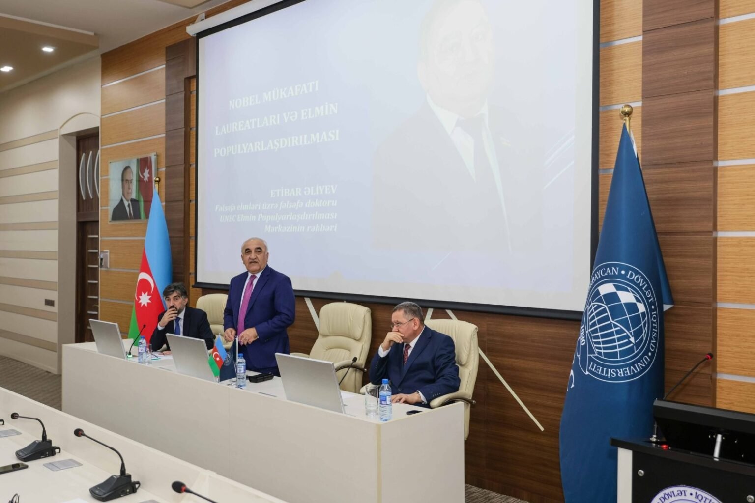 Выступление докладчика на тему Нобелевских лауреатов и популяризации науки в конференц-зале UNEC, на экране — презентация, на заднем плане — флаг Азербайджана