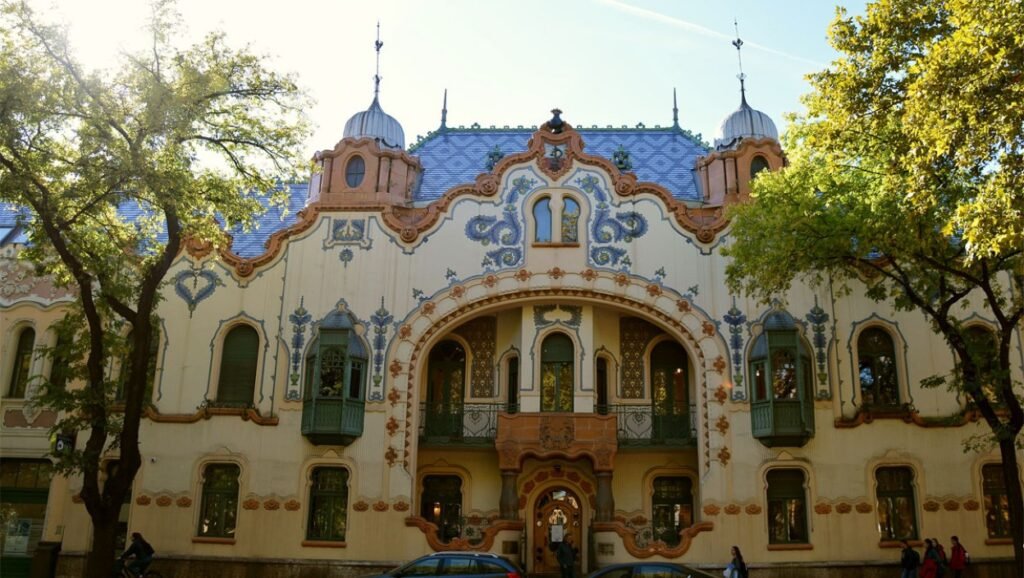 Фасад исторического дворца Райхле в центре Суботицы с синей черепичной крышей в стиле венгерского модерна, ажурной лепниной и изогнутыми балконами; по бокам фотографии — зелёные кроны деревьев, на тротуаре у входа — силуэты прохожих