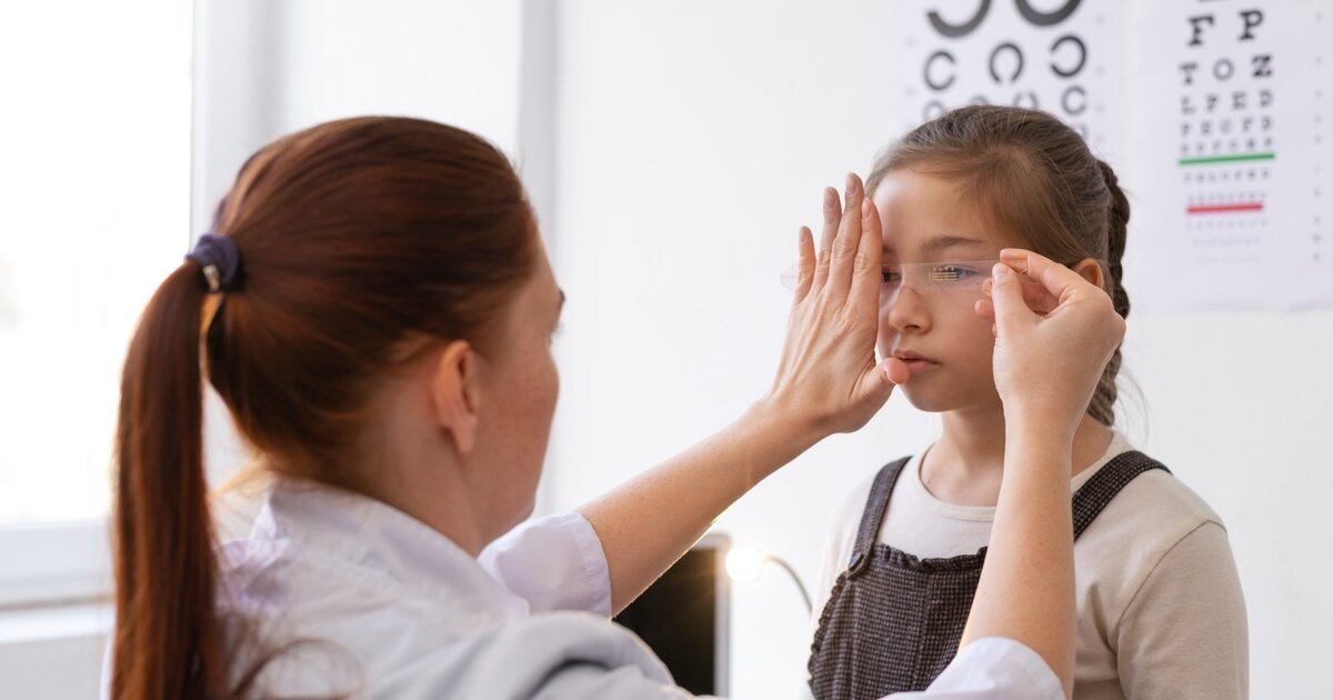 Врач подбирает очки девочке на фоне таблицы для проверки зрения в офтальмологическом кабинете
