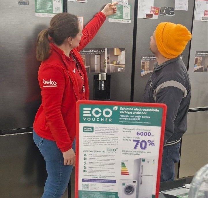 Покупательница выбирает холодильник в магазине бытовой техники в рамках программы EcoVoucher — рядом информационный стенд с условиями получения скидки до 70%