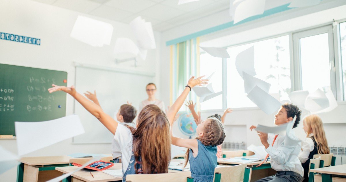 Дети радостно подбрасывают листы бумаги в классе на фоне школьной доски с задачами