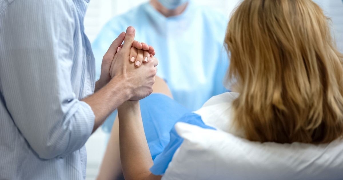 Роженица в родильном зале держит за руку близкого человека, рядом работает врач в голубом халате и маске. Кадр передаёт атмосферу родов в условиях медучреждения