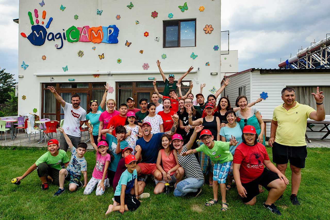 Волонтёры и подопечные Ассоциации Magic позируют у здания MagiCAMP с логотипом-ладошкой на фасаде