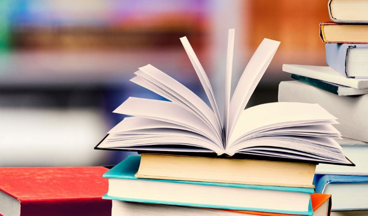 Раскрытый школьный учебник на стопке цветных книг на фоне библиотечных полок