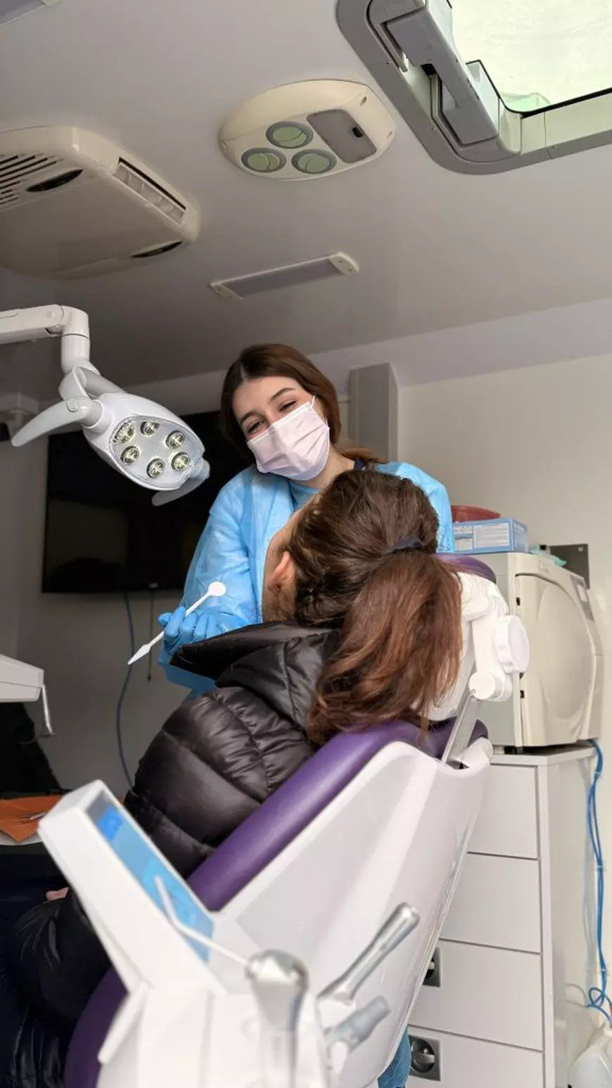 Молодой стоматолог в голубом халате и маске проводит бесплатный осмотр пациентки в мобильном стоматологическом кабинете Каравана здоровья полости рта