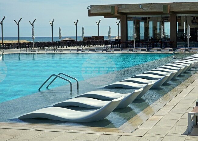 Открытый бассейн с рядом белых пластиковых шезлонгов и сложёнными зонтами на территории черноморского курорта накануне летнего сезона, за которым виднеется кромка Чёрного моря и терраса ресторана