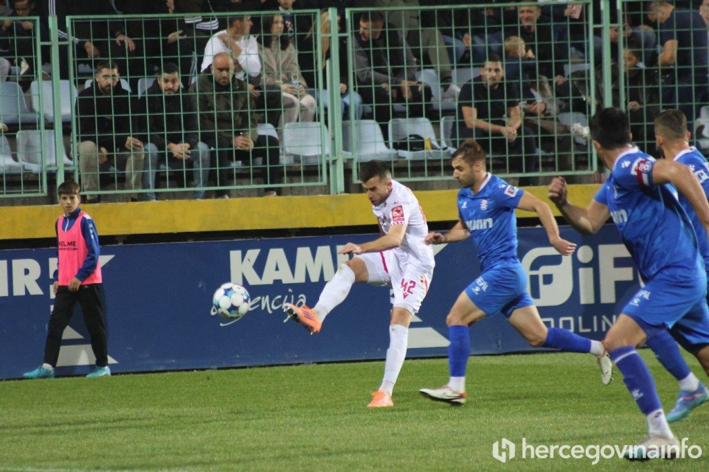 Игрок «Зриньски» в белой форме (№42) бьёт по воротам в окружении защитников «Широки Бриег» в синем — кадр из герцеговинского дерби, отсылка к матчу на стадионе Печара