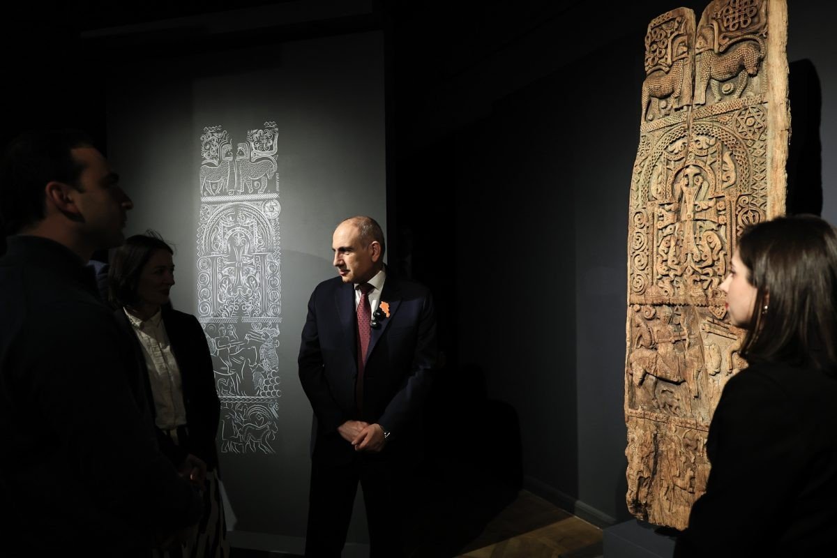 Премьер-министр Никол Пашинян и посетители осматривают резную деревянную дверную панель на открытии выставки, на стене за ними — проекция контурной схемы артефакта