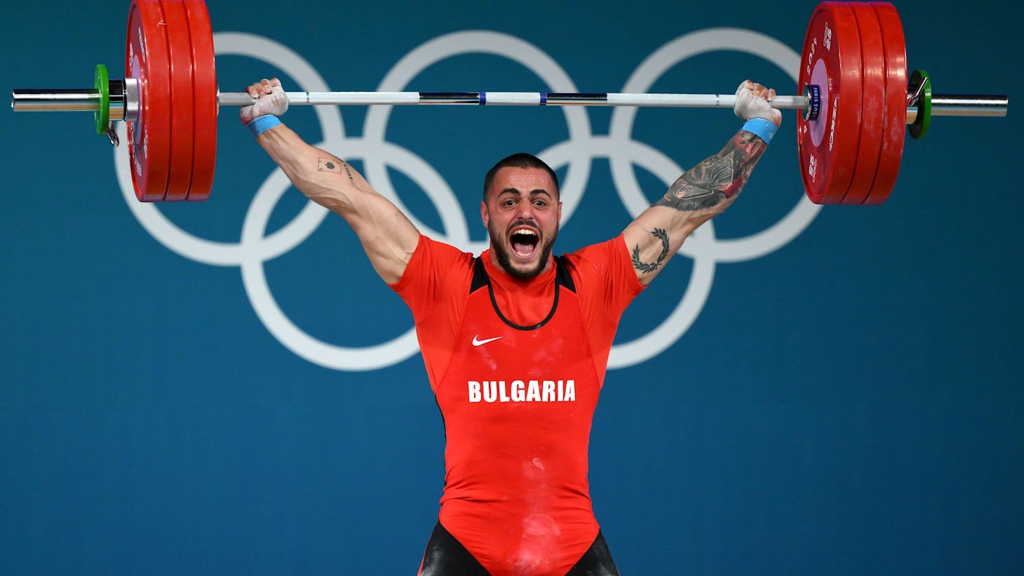 Болгарский тяжелоатлет Карлос Насар в красной форме сборной Болгарии фиксирует штангу над головой на фоне олимпийских колец