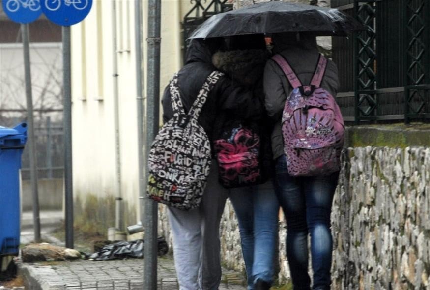 Школьницы с рюкзаками укрываются под зонтом от дождя на улице греческого города