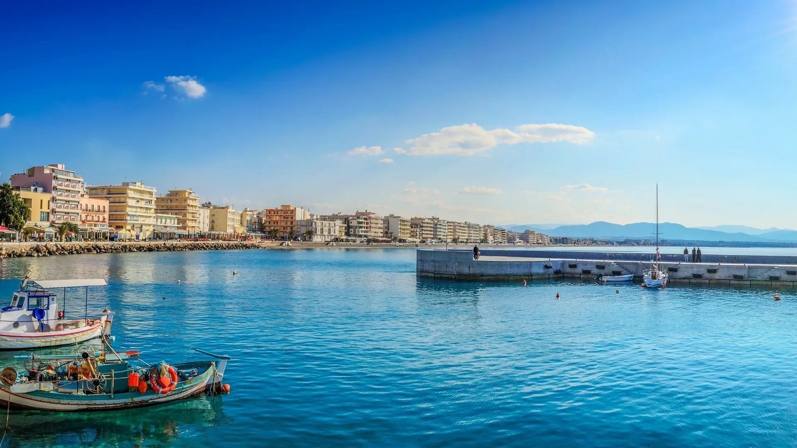 Набережная Лутраки — рыбацкие лодки в лазурной воде порта и панорама прибрежных зданий на фоне гор