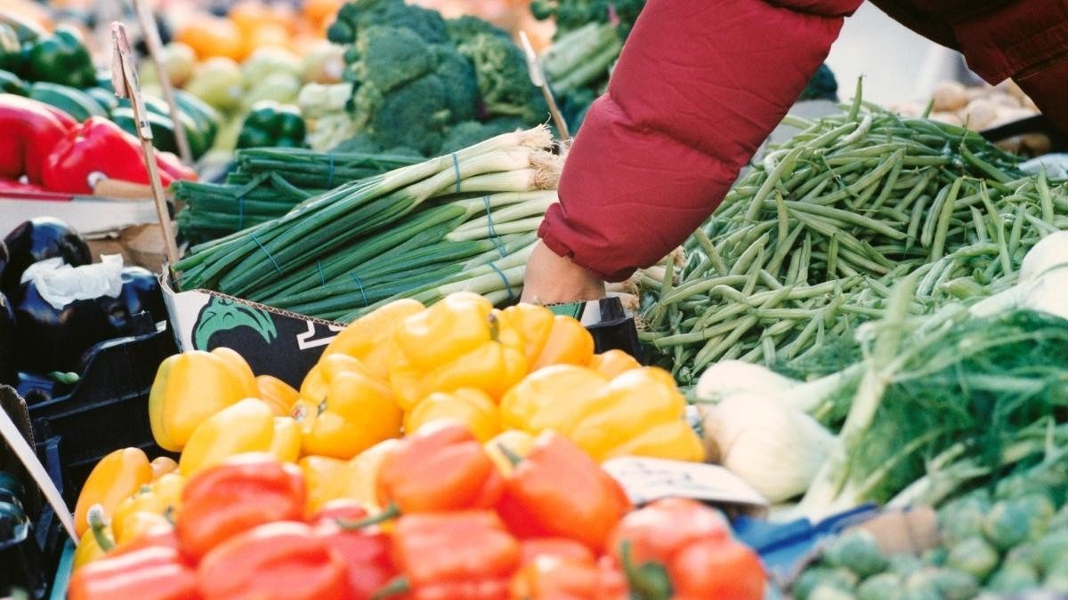 Покупатель выбирает зелёный лук у лотка с перцем, стручковой фасолью и другими овощами на румынском рынке