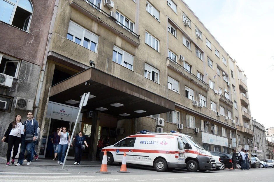 Главный вход в белградский роддом ГАК «Народни фронт»: у подъезда с логотипом клиники припаркован реанимобиль с надписью AMBULANCE, из здания выходят посетители