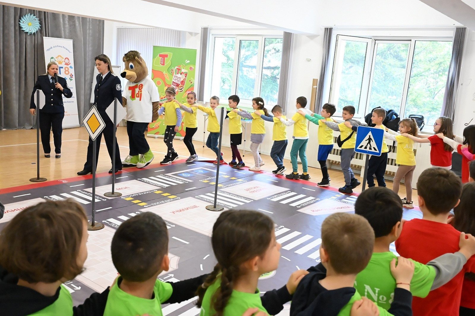 Дети из подготовительной группы цепочкой следуют за сотрудницей полиции и талисманом TEDI по интерактивному коврику с пешеходными переходами и дорожными знаками в зале детского сада