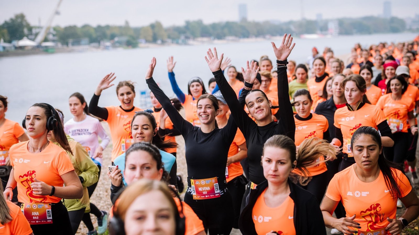 Участницы женского забега в оранжевых футболках Nike приветствуют камеру на дистанции вдоль реки Сава