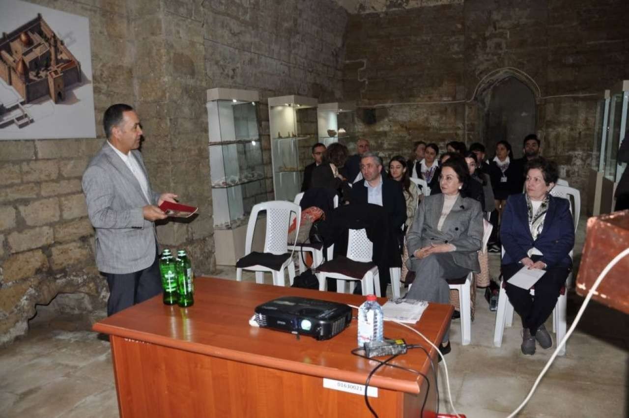 Эксперт проводит презентацию о реставрации «Пир Хусейн Ханегях» в старинном каменном зале заповедника: слева — архитектурная схема памятника, в витринах — археологические находки