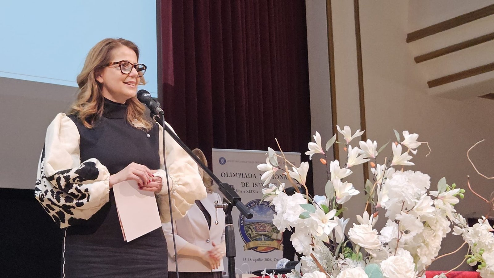 Ведущая церемонии выступает на трибуне рядом с баннером национальной олимпиады по истории в Ораде