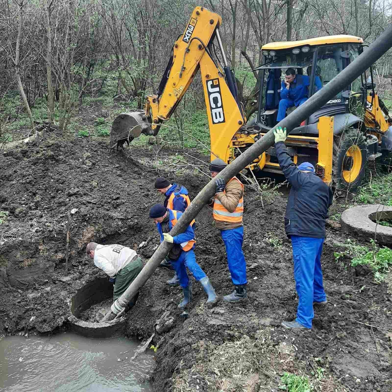 Работники S.A. „Apă-Canal Chișinău” в синей спецодежде протягивают новую трубу через размытый котлован в лесополосе — экскаватор JCB придерживает плеть магистрального трубопровода