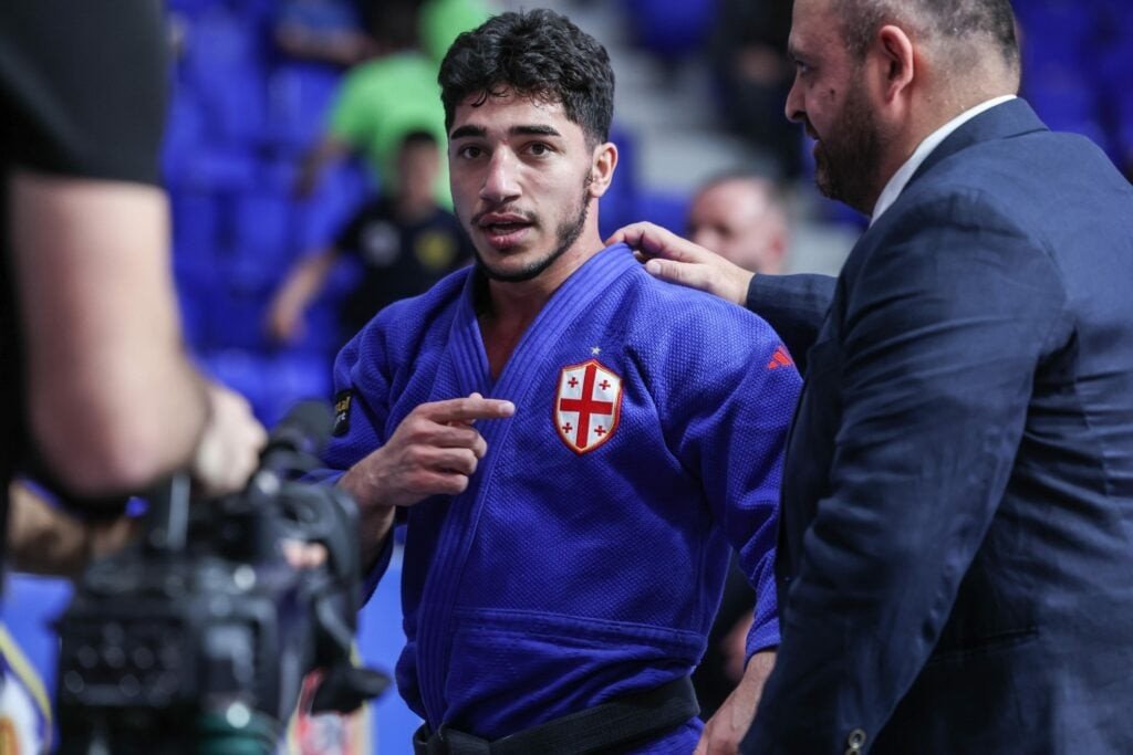 Грузинский дзюдоист в сине-белом кимоно с крестом святого Георгия беседует с тренером у края татами: хозяева чемпионата Европы 2026 рассчитывают на домашнюю поддержку зала