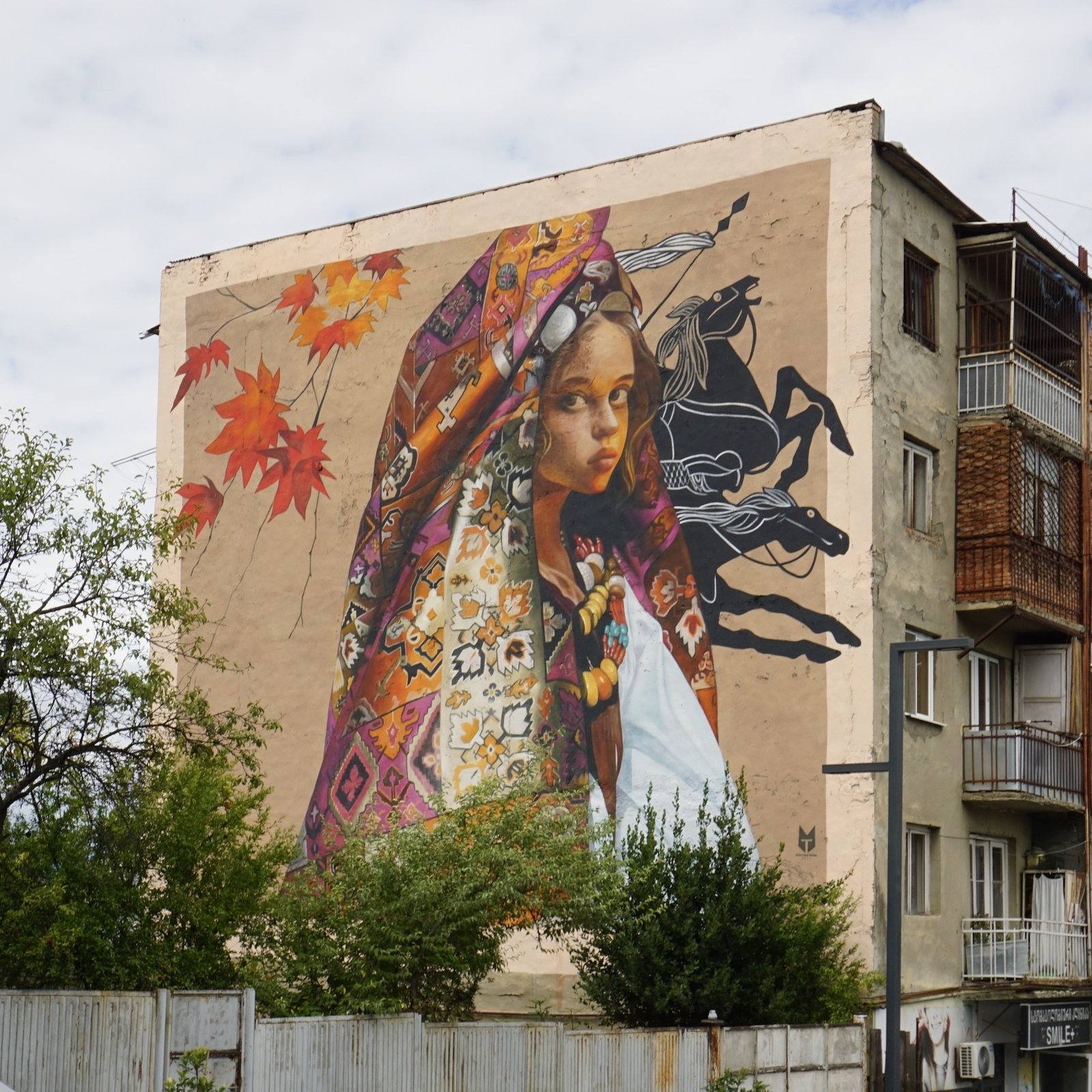 Мурал на стене дома в Тбилиси: девочка в традиционной грузинской шали с орнаментом и осенними листьями