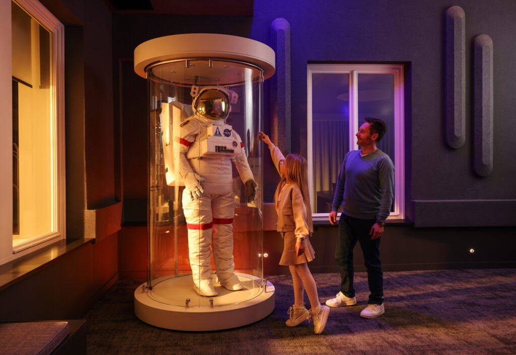 Отец с дочерью разглядывают подлинный скафандр NASA в стеклянной витрине-цилиндре на экспозиции Палаты науки — отсылка к программе «Аполлон»