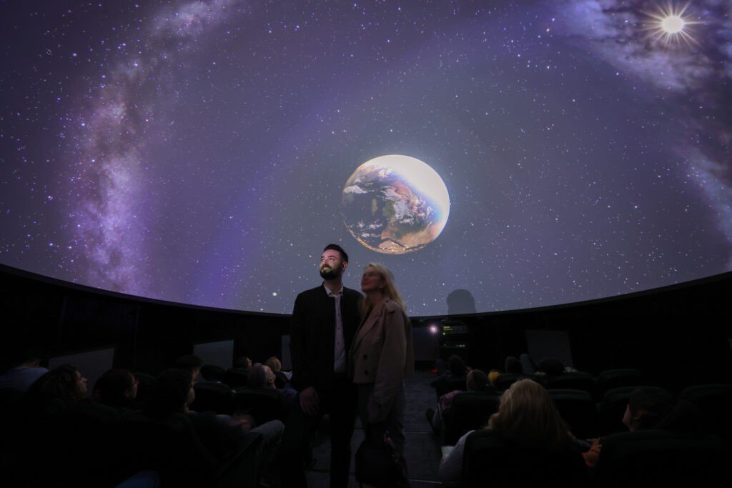 Сеанс в планетарии Палаты науки: на куполе проецируются Земля, Млечный Путь и россыпь звёзд, зрители в креслах и двое посетителей смотрят вверх