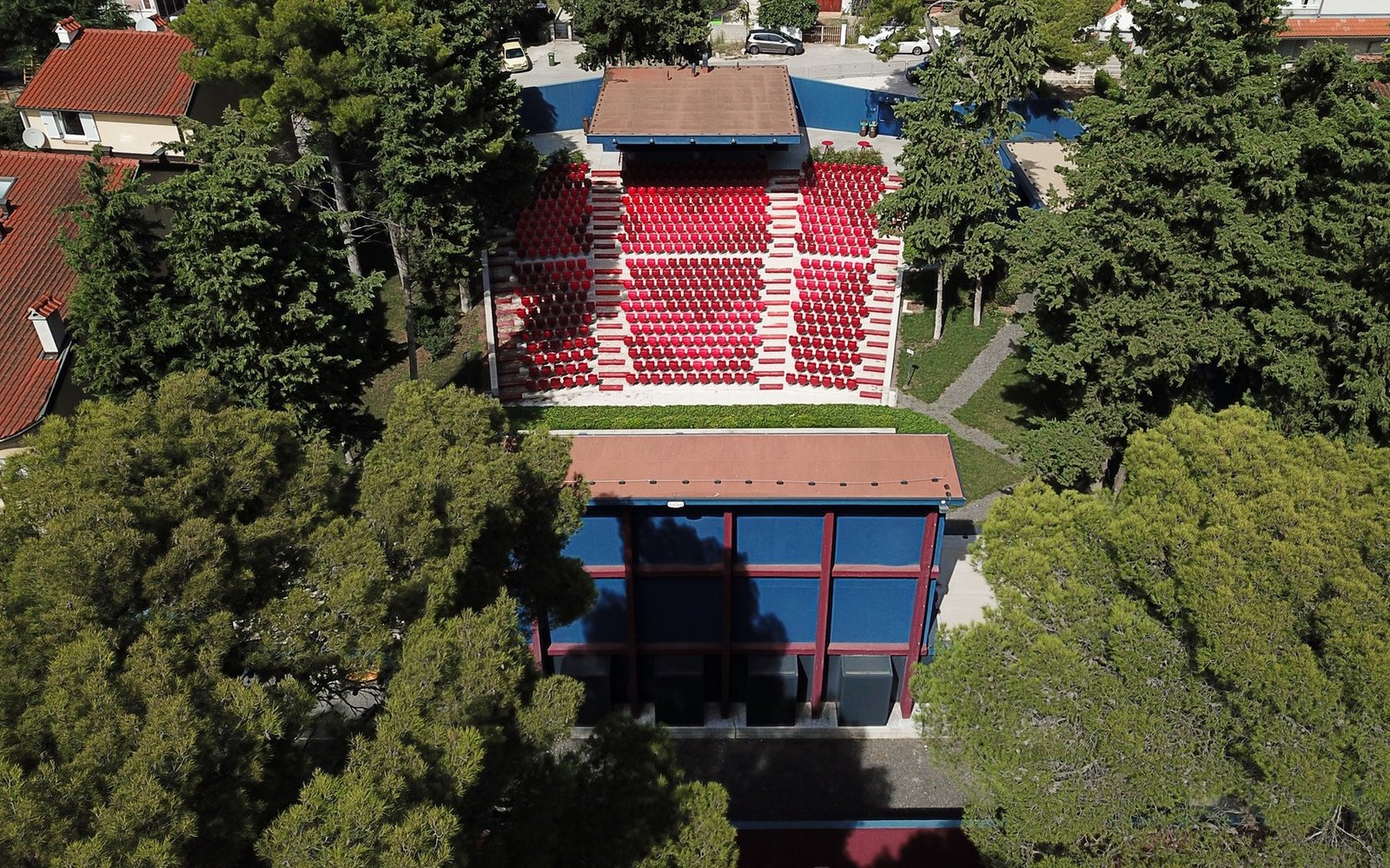 Летняя сцена с красно-белыми рядами зрительских мест и павильоном среди высоких сосен, вид сверху