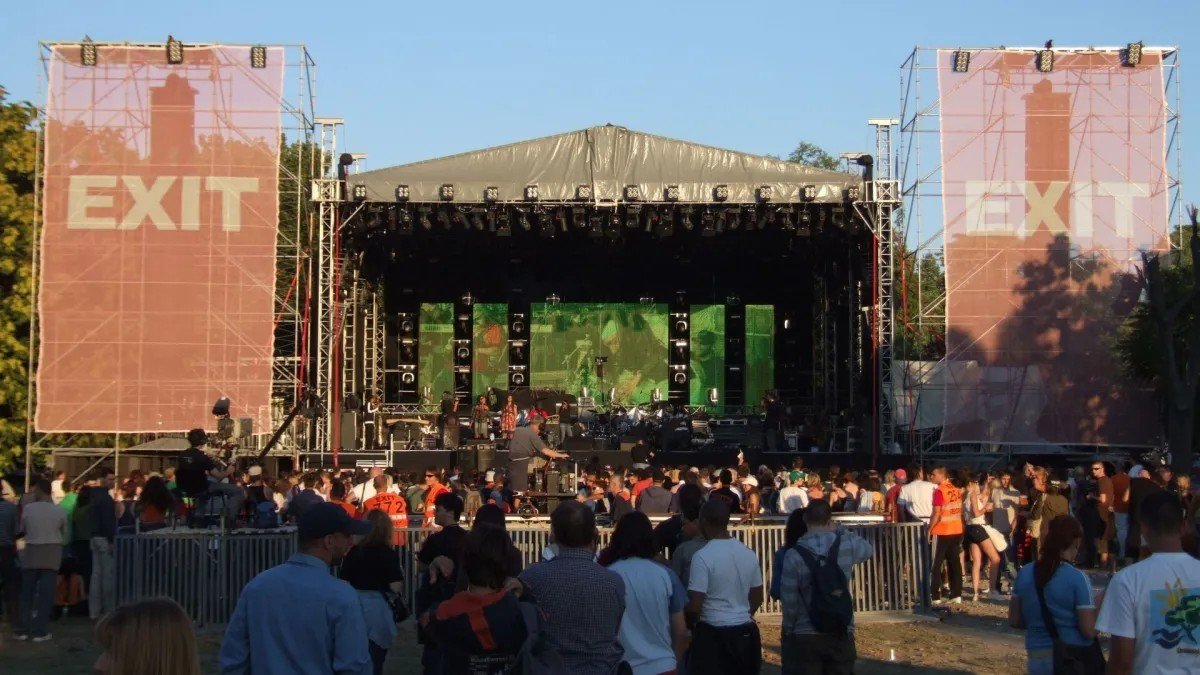Главная сцена фестиваля EXIT в дневное время с большими баннерами EXIT, сценическим оборудованием и посетителями фестиваля на переднем плане