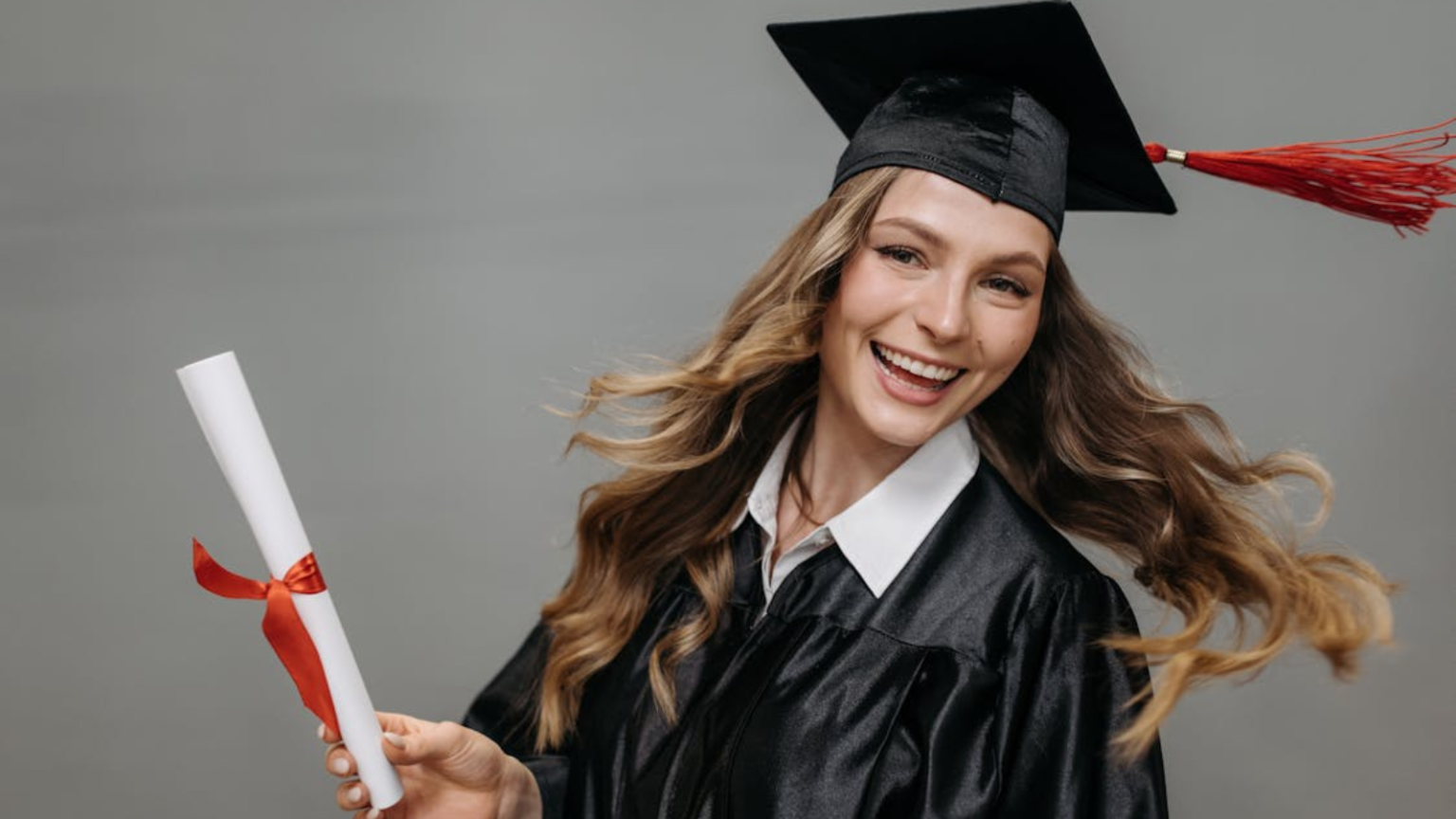 Улыбающаяся девушка в чёрной академической мантии и квадратной шапочке-конфедератке держит в руке свёрнутый диплом, перевязанный красной лентой