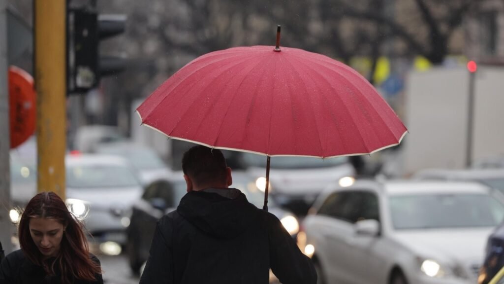 Дожди и холод в Болгарии: апрель начался с непогоды Прохожий с красным зонтом идёт под дождём по городской улице на фоне автомобилей и светофоров