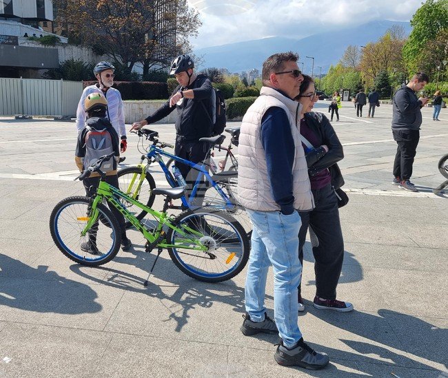 Велосипедисты и прохожие на солнечной площади в Софии на фоне зелёных деревьев и склона Витоши