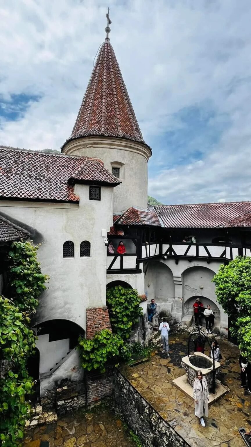 Островерхая башня замка Бран с красной черепичной крышей над побелёнными стенами внутреннего двора и туристами у каменного колодца