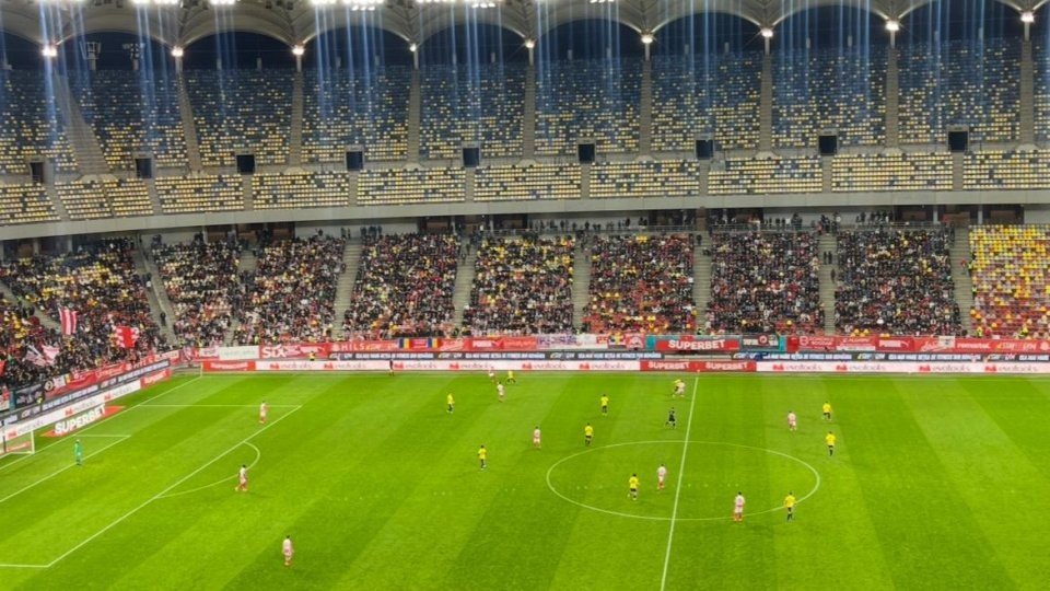 Общий план матча на бухарестской Arena Națională: игроки «Динамо» в красно-белой форме атакуют оборону «Университати Клуж» в жёлтом, трибуны заполнены болельщиками со скарфами и баннерами