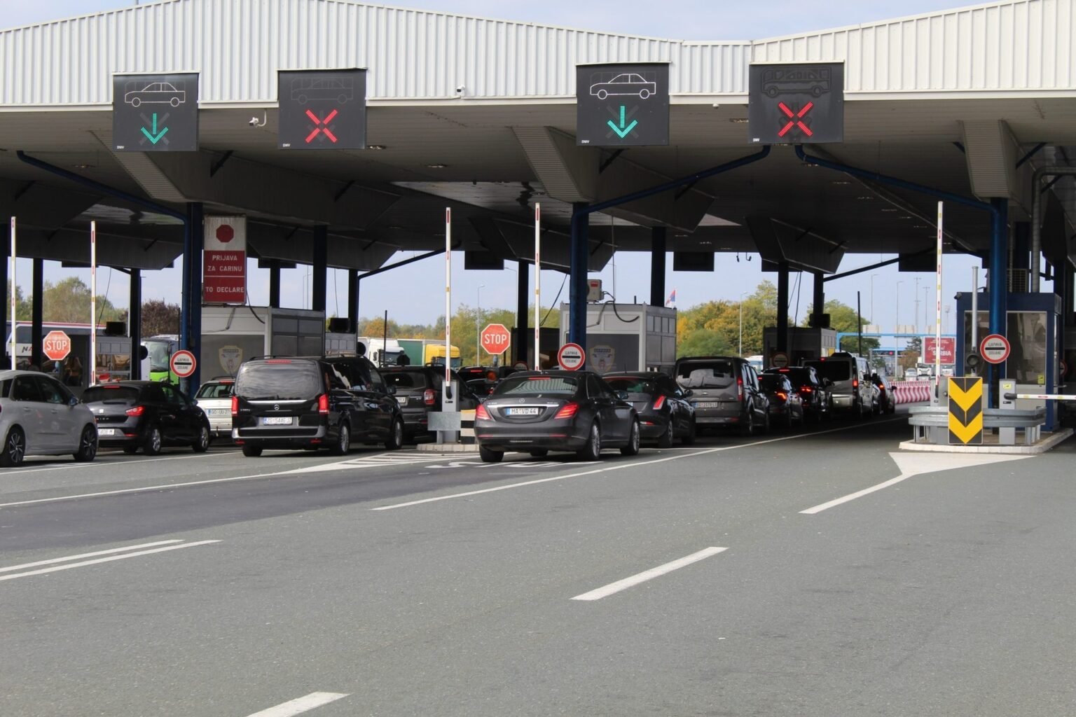 Очередь легковых автомобилей под навесом сербского пограничного пункта: водители ждут паспортного контроля у автоматических шлагбаумов и табло с символами «въезд/выезд»