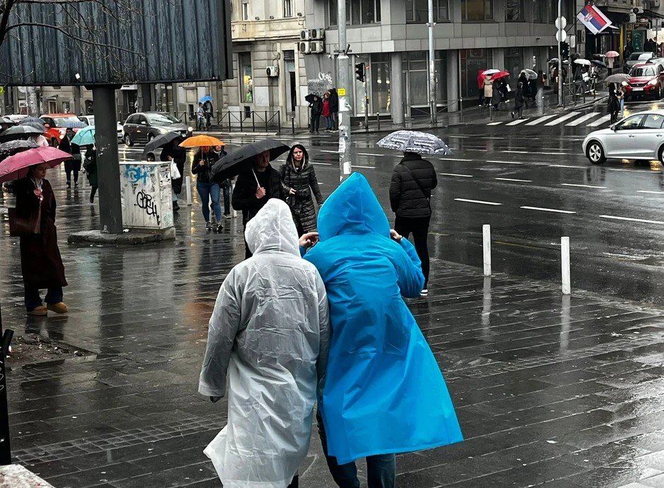 Пешеходы в дождевиках и с зонтами переходят мокрый перекрёсток в Белграде во время сильного дождя