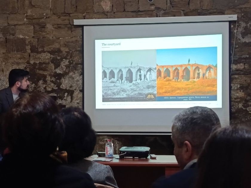 Участники конференции в каменном зале рассматривают слайд "The courtyard" со сравнением исторического и современного видов двора памятника архитектуры