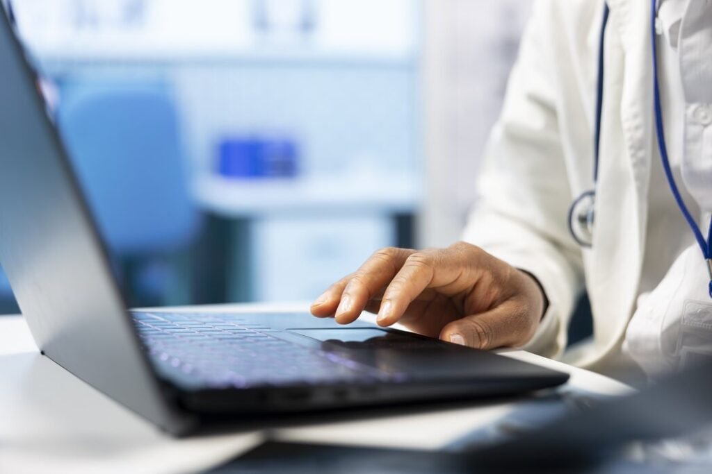 Сербия перешла на электронную подачу заявлений на возмещение зарплаты Врач в белом халате со стетоскопом работает за ноутбуком в медицинском кабинете