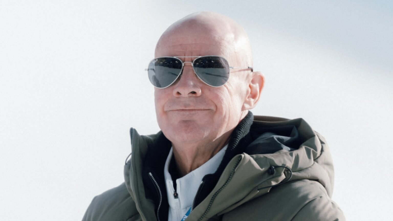 Президент FIS Йохан Элиаш в тёмных очках-авиаторах и оливковой зимней куртке на фоне заснеженного склона