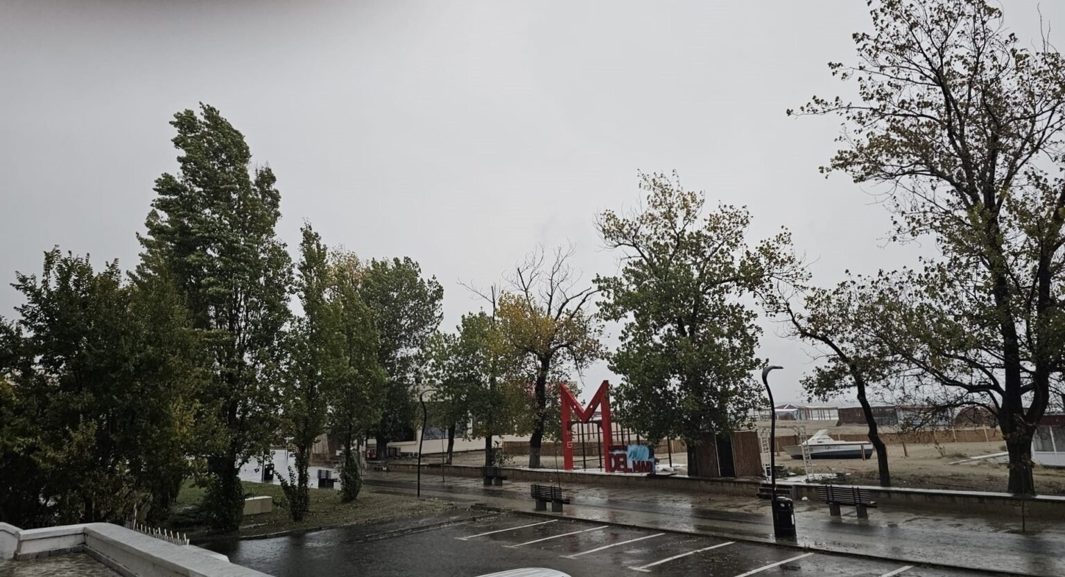 Пустая мокрая набережная и гнущиеся под ветром деревья у арт-объекта «Mamaia» на фоне серого неба — иллюстрация холодной и закрытой погоды на румынском побережье