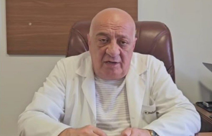 Пожилой врач в белом халате сидит за рабочим столом в кабинете поликлиники Бюрегавана