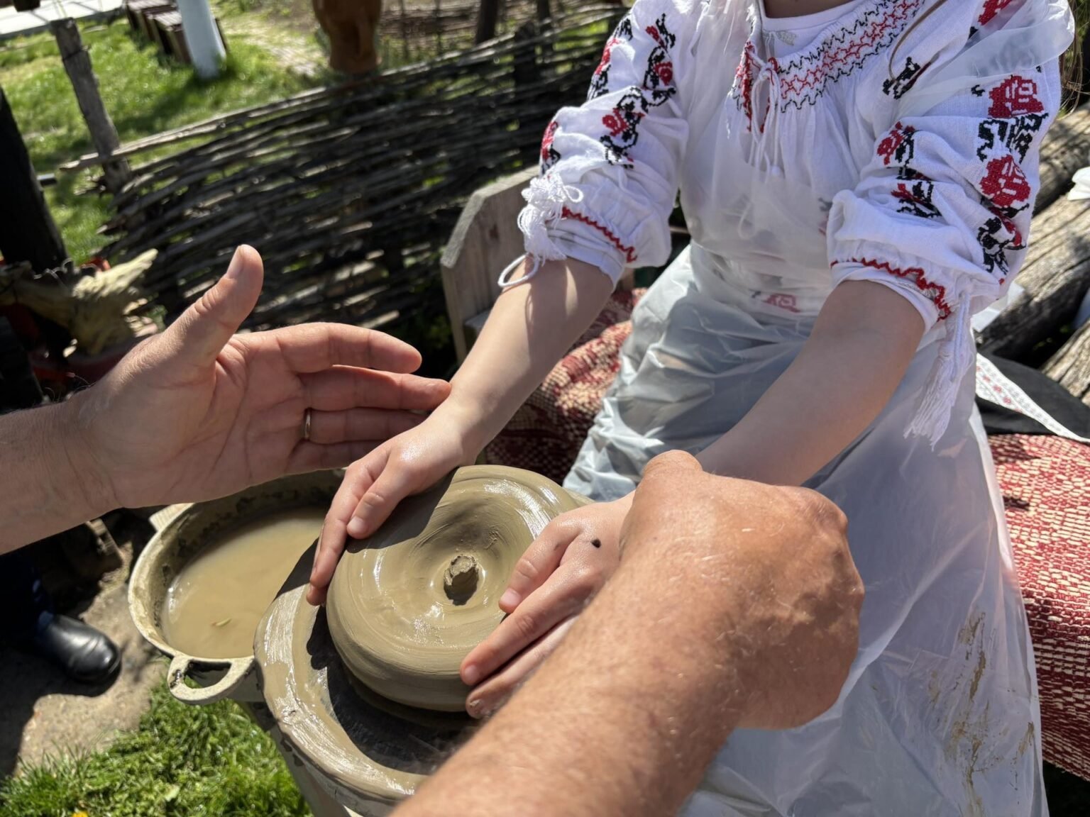 Мастер обучает участницу в традиционной вышитой рубашке гончарному ремеслу на гончарном круге во время мастер-класса проекта KULtura