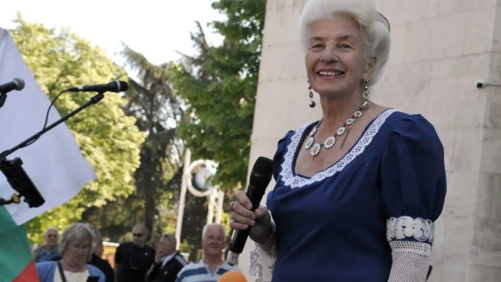 Янка Рупкина с микрофоном в тёмно-синем нарядном платье на открытой площадке, рядом болгарский флаг и публика