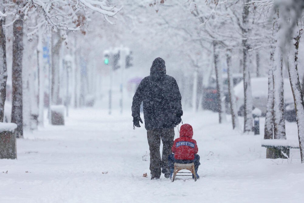 Взрослый везёт ребёнка на санках по заснеженной аллее во время обильного снегопада