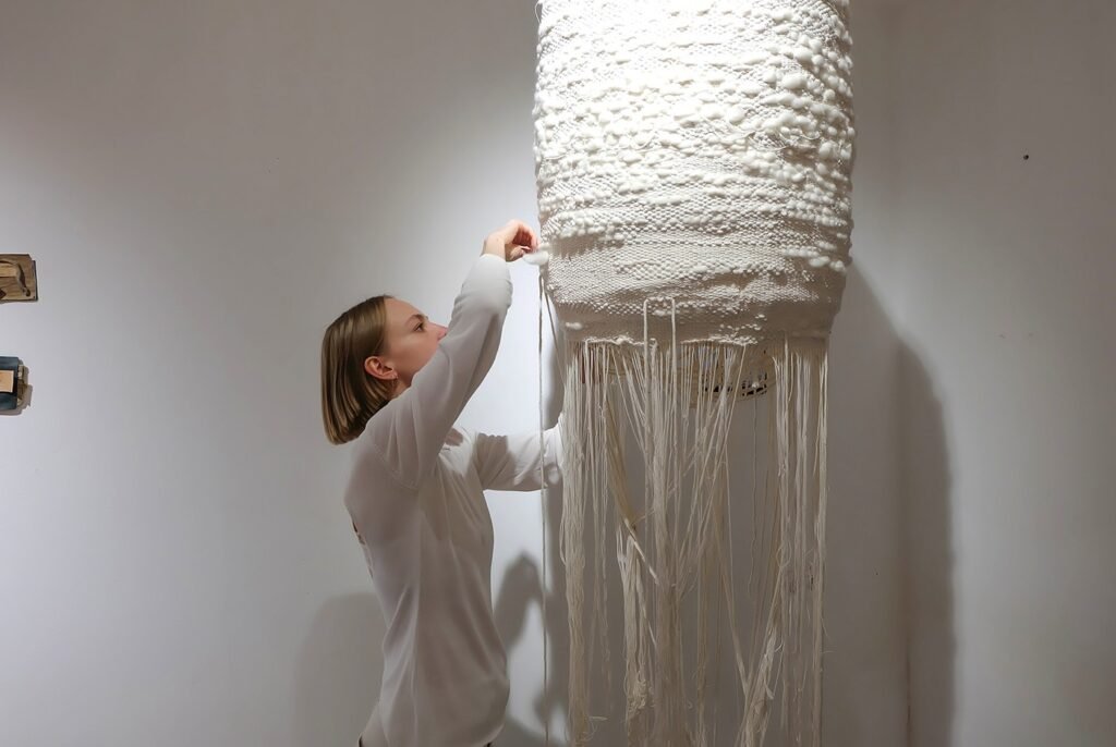 Художница поправляет подвесную тканую скульптуру с длинными нитевыми волокнами в экспозиции Soft Structures в Тбилиси