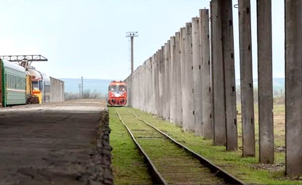 Красно-белый дизель-локомотив подходит к пограничной станции на восстановленном участке Прут — Фэлчиу; слева на запасном пути стоят старые пассажирские вагоны, справа вдоль колеи тянется бетонный забор