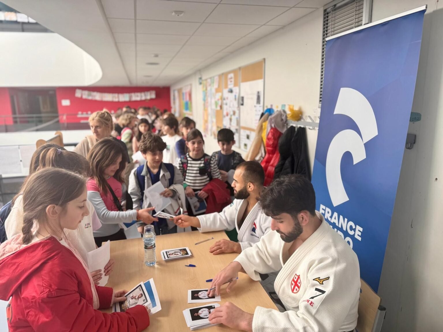 Ученики Французской школы Кавказа в Тбилиси окружили стол, за которым грузинские дзюдоисты в белых кимоно подписывают карточки для юных поклонников; рядом развёрнут баннер France Judo