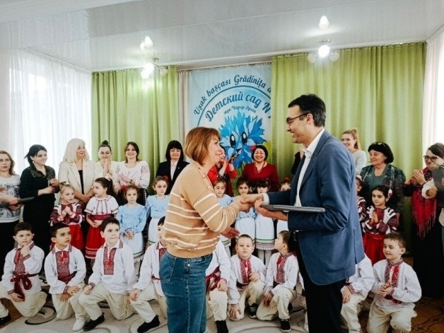 Женщина передает ноутбук мужчине в костюме на фоне детей в национальных костюмах