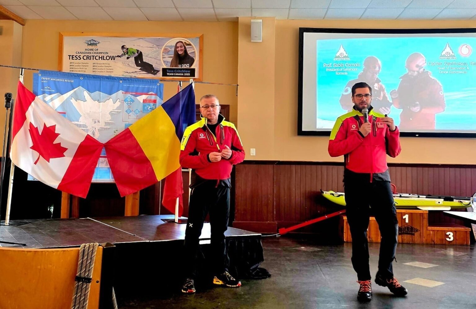 Два спасателя Salvamont Romania в красных куртках стоят на сцене конференц-зала. За ними — флаги Канады и Румынии, на экране транслируется презентация. Один из спасателей держит микрофон