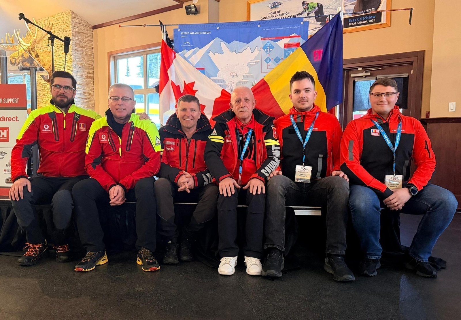 Шесть членов румынской делегации Salvamont сидят на скамье в помещении конференции. За ними видны флаги Канады и Румынии, баннер конгресса. Спасатели одеты в красные и чёрные форменные куртки, на шеях — бейджи участников