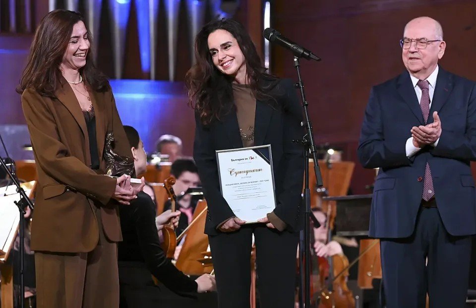 Музыка как социальная миссия: Fest Team запускает четыре программы помощи в Пловдиве Церемония награждения на сцене — женщина получает сертификат на фоне симфонического оркестра